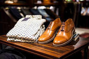 Brown leather dress shoes and folded patterned shirts are displayed on a wooden table in a clothing store.
